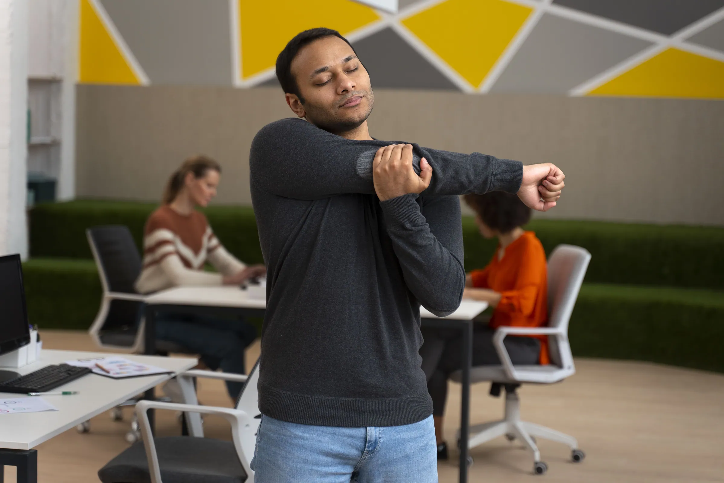 Office worker standing by his desk stretching his arm across his chest while coworkers work in the background
